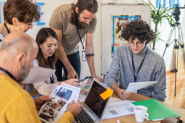 Five casually dressed media agency colleagues of different genders and ethnicities gather around a desk examining papers and photographs. There is a camera on a tripod in the corner